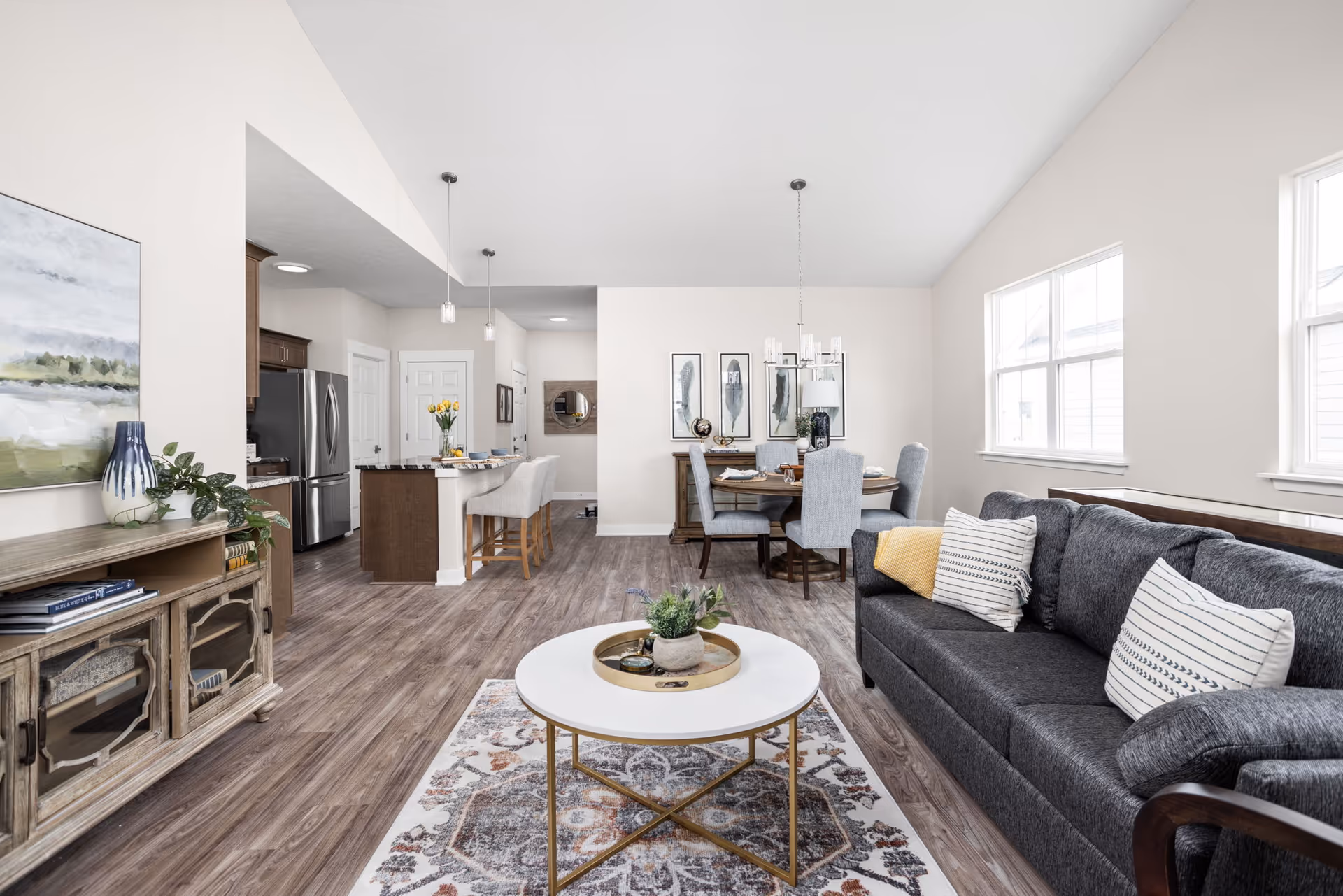 A bright and spacious open-concept living area featuring a dark gray sofa with striped pillows, a round white coffee table with a decorative tray and plant, a patterned area rug, a wooden sideboard with books and a vase, a dining table with six upholstered chairs, and a kitchen with stainless steel appliances and a breakfast bar with two stools. The room has large windows allowing natural light to fill the space.