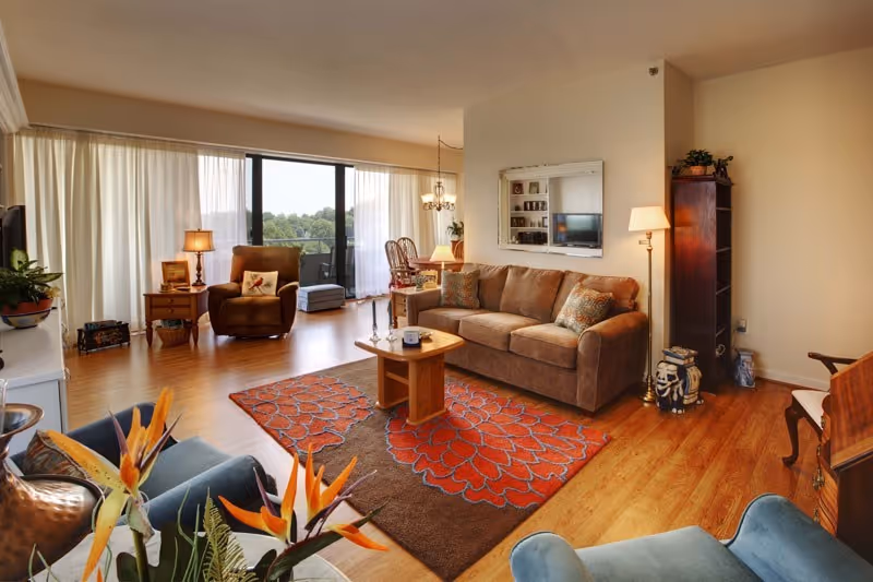 A cozy living room with a brown sofa, a wooden coffee table on a red and brown patterned rug, two armchairs, a floor lamp, and a large window with sheer curtains letting in natural light. There is a bookshelf and a TV mounted on the wall.