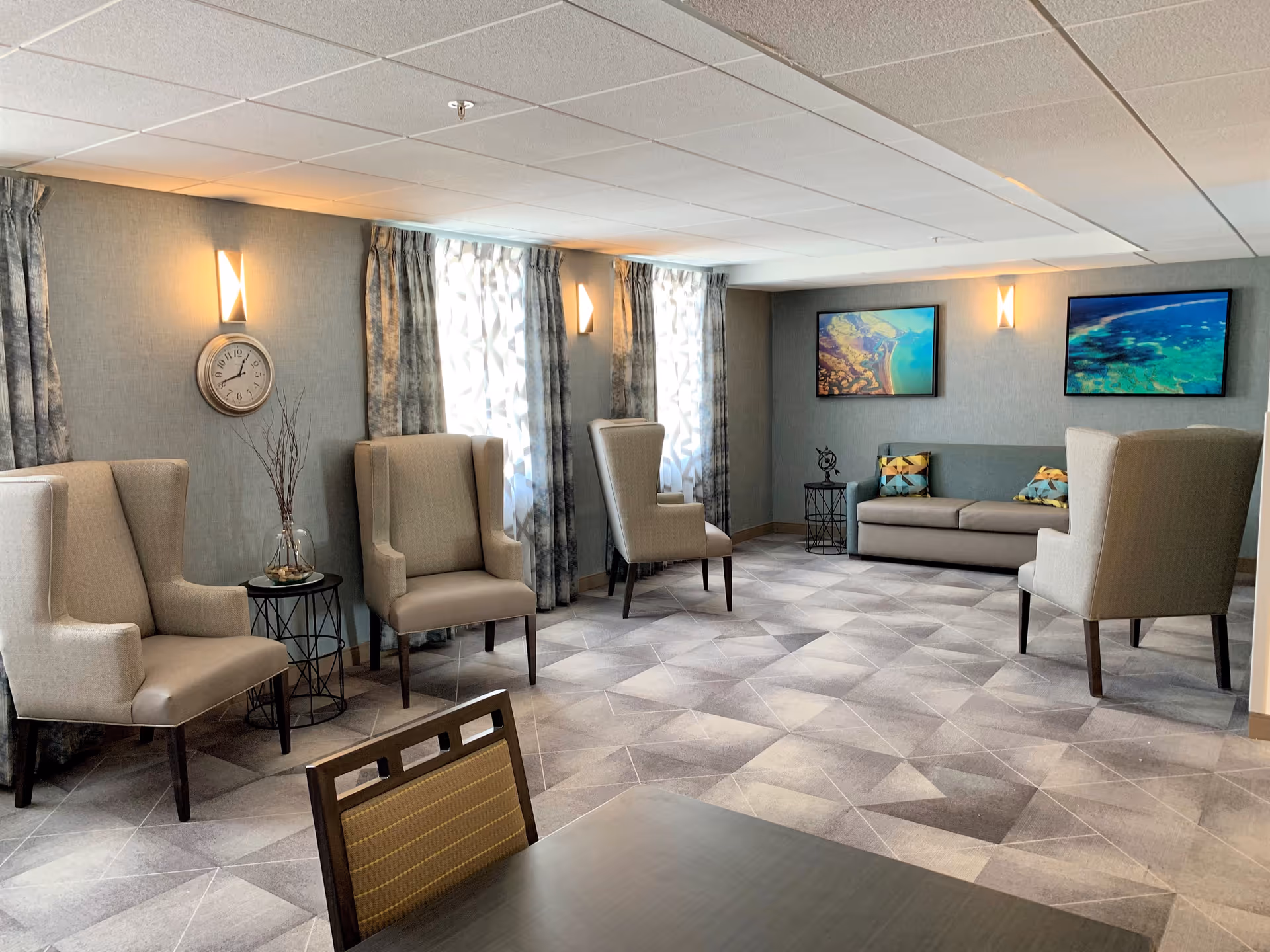 Well-lit sitting room with upholstered wingback chairs, a sofa, side tables, wall art and patterned carpet.