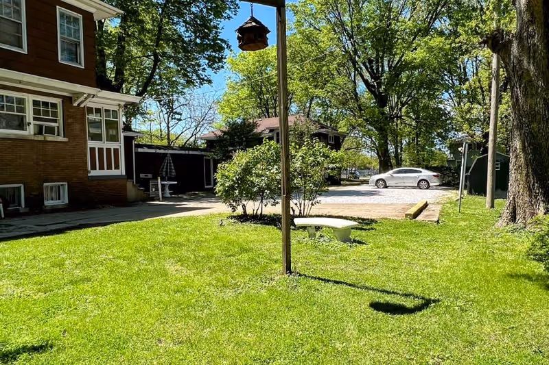 A sunny outdoor area at a senior living facility featuring a green lawn, a bird feeder hanging from a pole, a concrete bench, bushes, trees, a parked white car, and parts of brick buildings in the background.