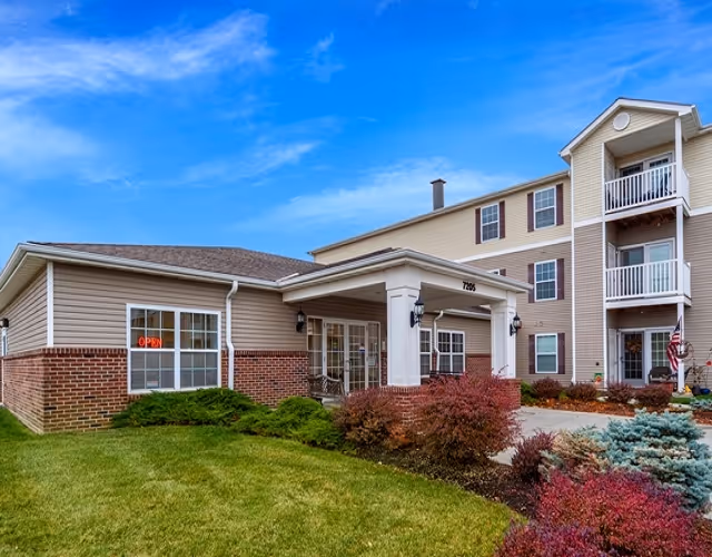 Exterior view of a multi-story senior living facility with beige siding and brick accents, featuring a covered entrance with white columns, well-maintained landscaping with green grass, shrubs, and colorful bushes under a blue sky.