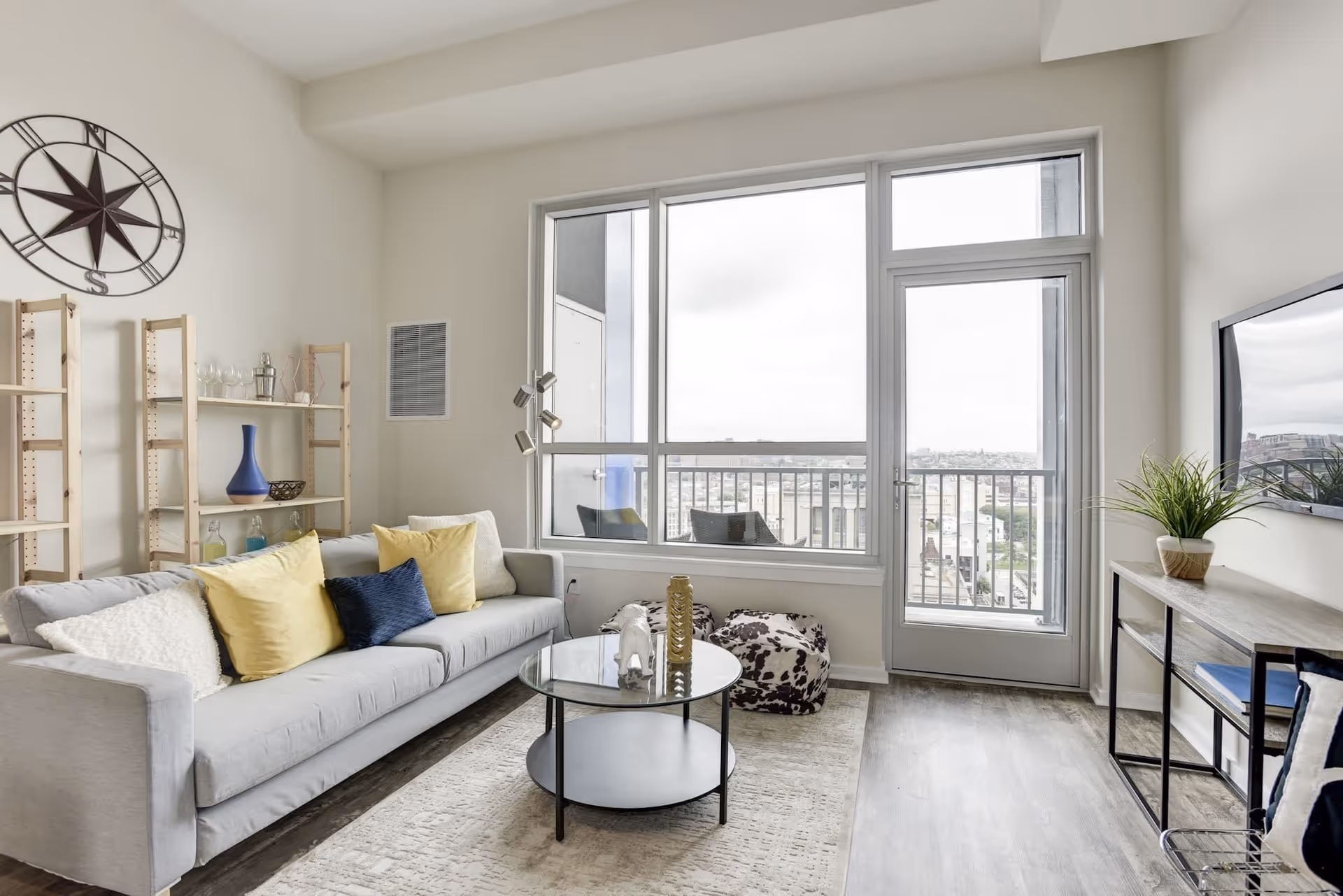 Bright modern living room with a gray sofa, colorful pillows, a round glass coffee table, shelving, and a large windowed door opening to a balcony.