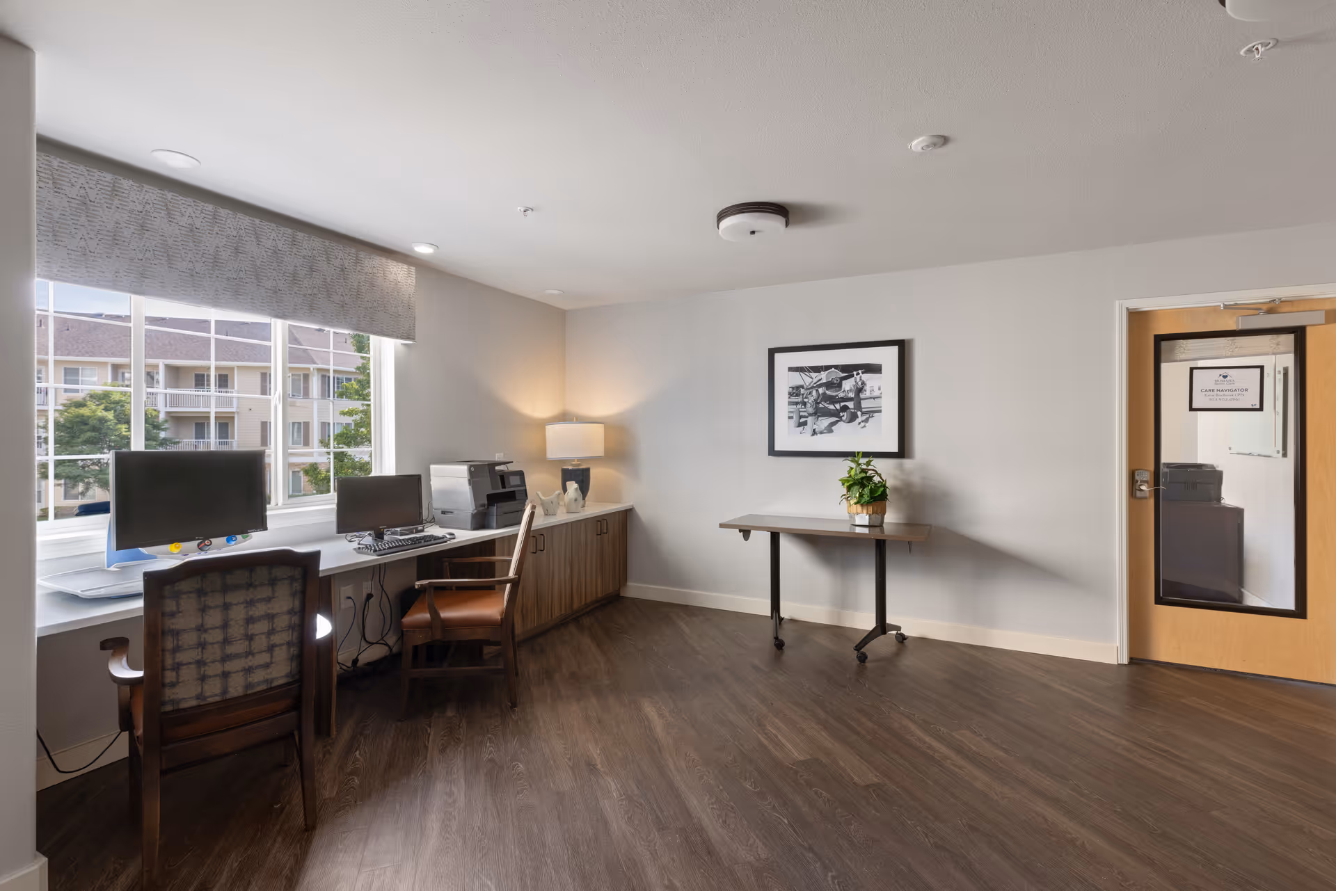 A bright room with two computer workstations by a large window, a wooden floor, a table with a potted plant against the wall, a framed black and white photograph, and a door with a window labeled 'Care Navigation'.