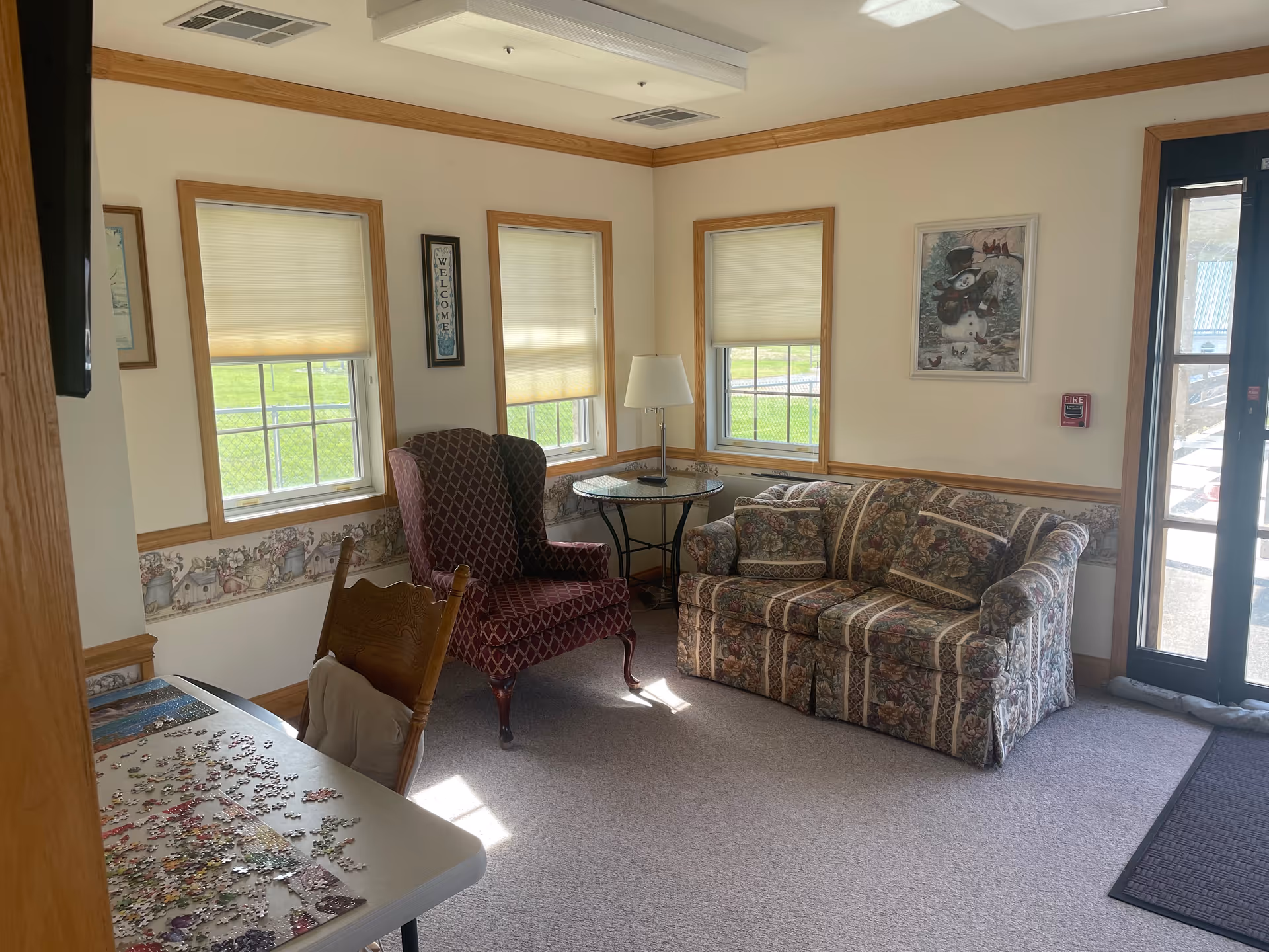 A cozy sitting area in an assisted living facility with a patterned armchair, a floral-patterned sofa, a round glass-top side table with a lamp, three windows with blinds partially drawn, a framed picture on the wall, and a table with a partially completed jigsaw puzzle in the foreground. The room has beige walls with a decorative wallpaper border and a glass door leading outside.
