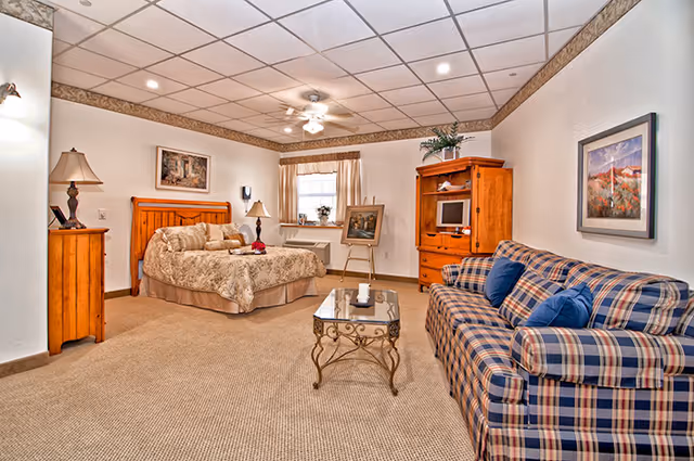 Spacious assisted-living bedroom suite with a bed, plaid sofa, coffee table, and wooden armoire under recessed ceiling lights.