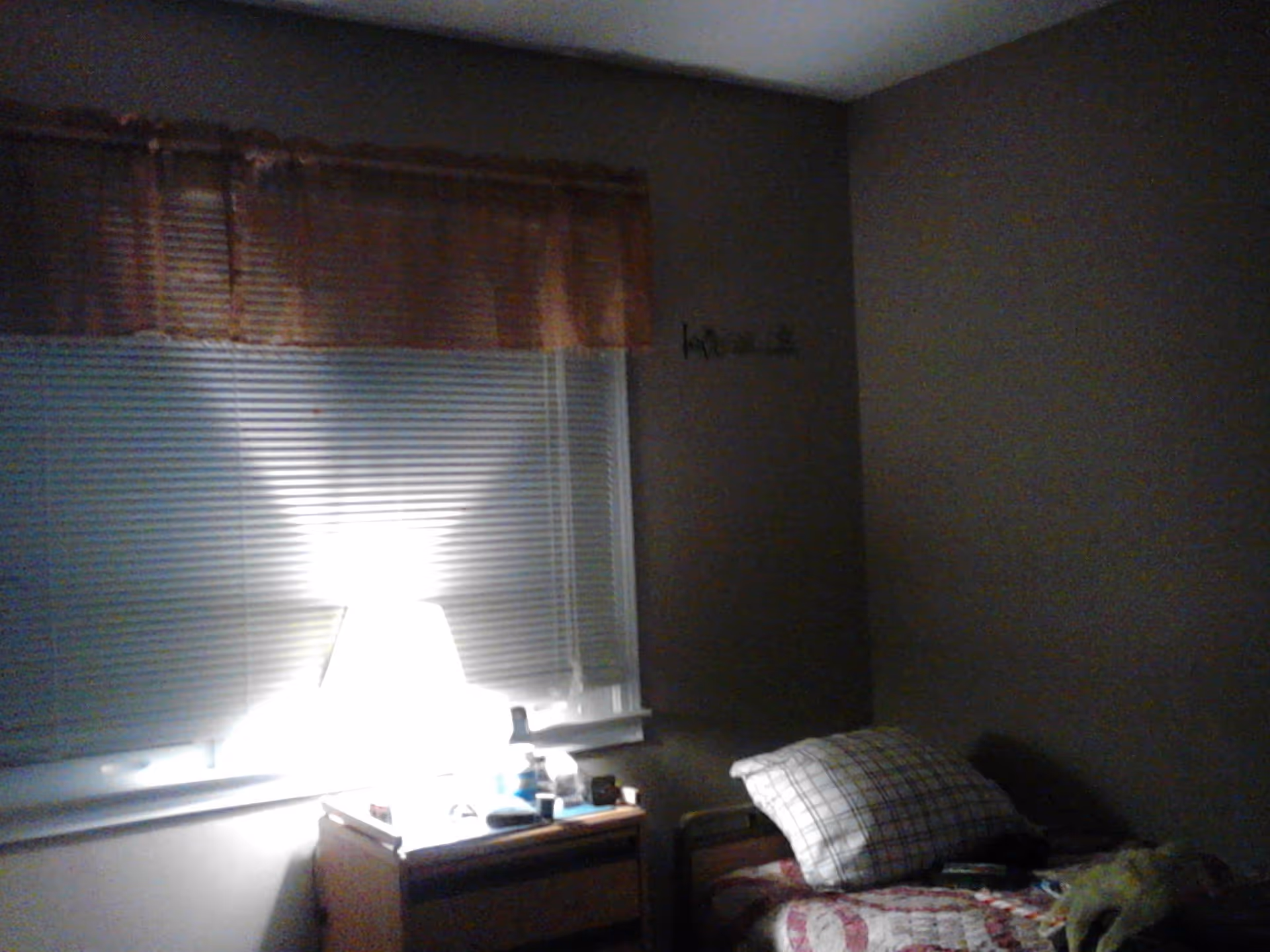 A dimly lit bedroom with a single bed covered in a patterned quilt and a plaid pillow. Next to the bed is a wooden nightstand with a bright lamp, various small items, and a window with closed blinds and a brown valance above it.