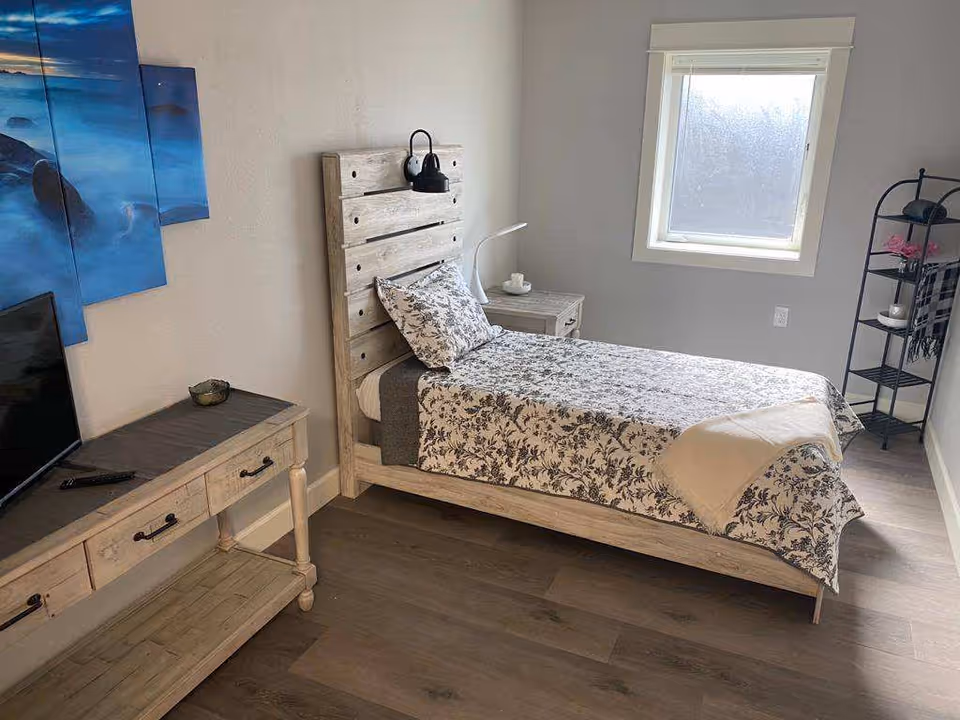 A small, neatly arranged bedroom with a single bed featuring a wooden headboard and floral bedding. There is a wooden nightstand with a lamp and a cup, a window with frosted glass, a wooden console table with a TV and remote, and a black metal shelving unit with decorative items and a folded blanket.