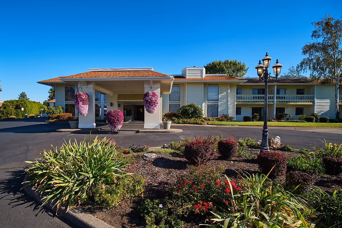 Front exterior view of Northridge Senior Living facility with a covered entrance, hanging flower baskets, a landscaped garden with various plants and flowers, and a clear blue sky.