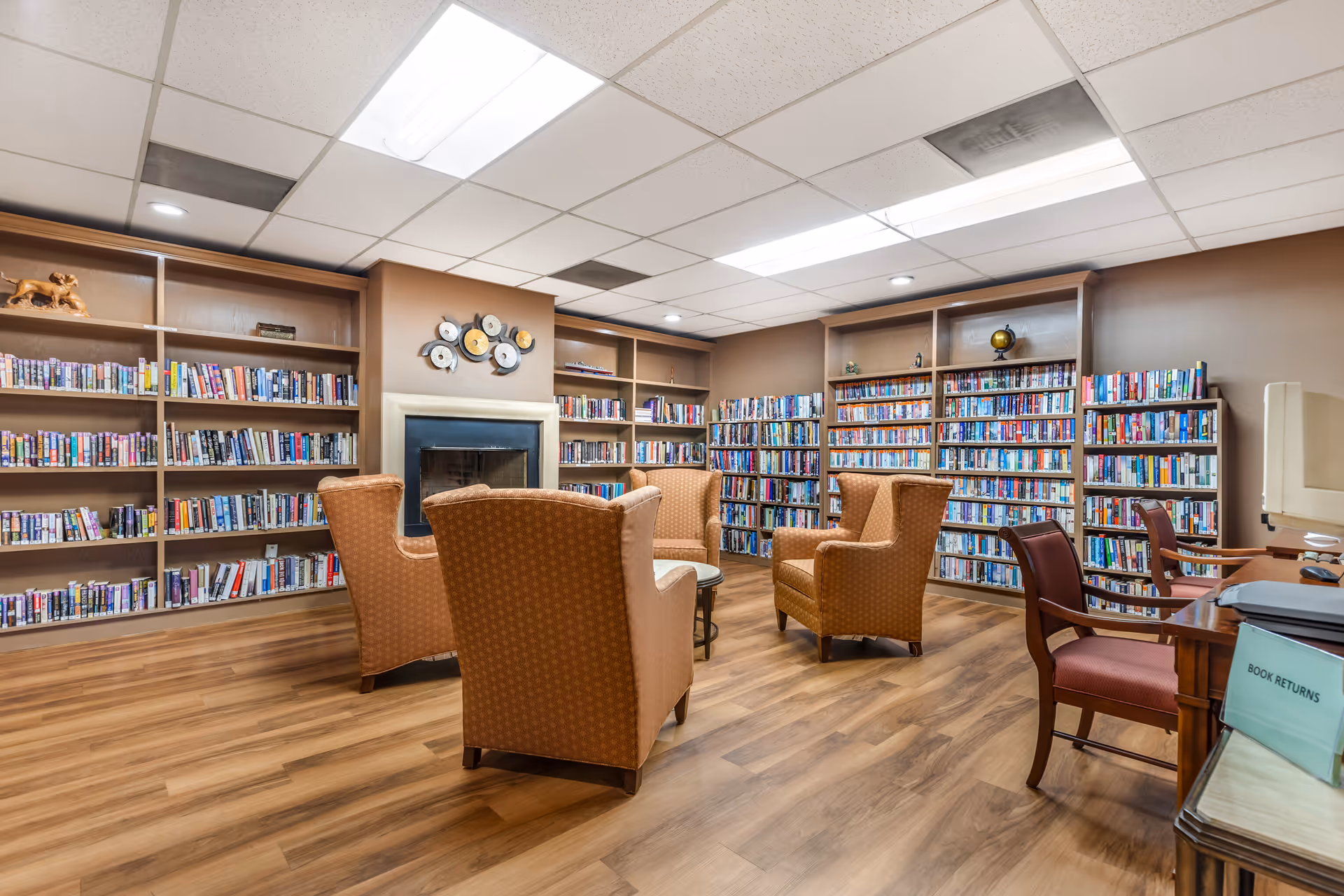 Library lounge with wall-to-wall bookshelves, several upholstered armchairs around a central table, and a fireplace.