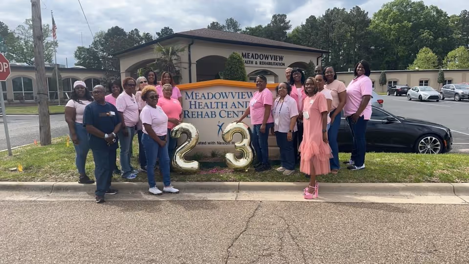 A group of people standing outside Meadowview Health and Rehab Center, posing in front of the facility's sign. They are wearing pink and casual clothing, and there are large gold balloons shaped as the numbers 2 and 3 in front of the sign. The building and parking lot are visible in the background.