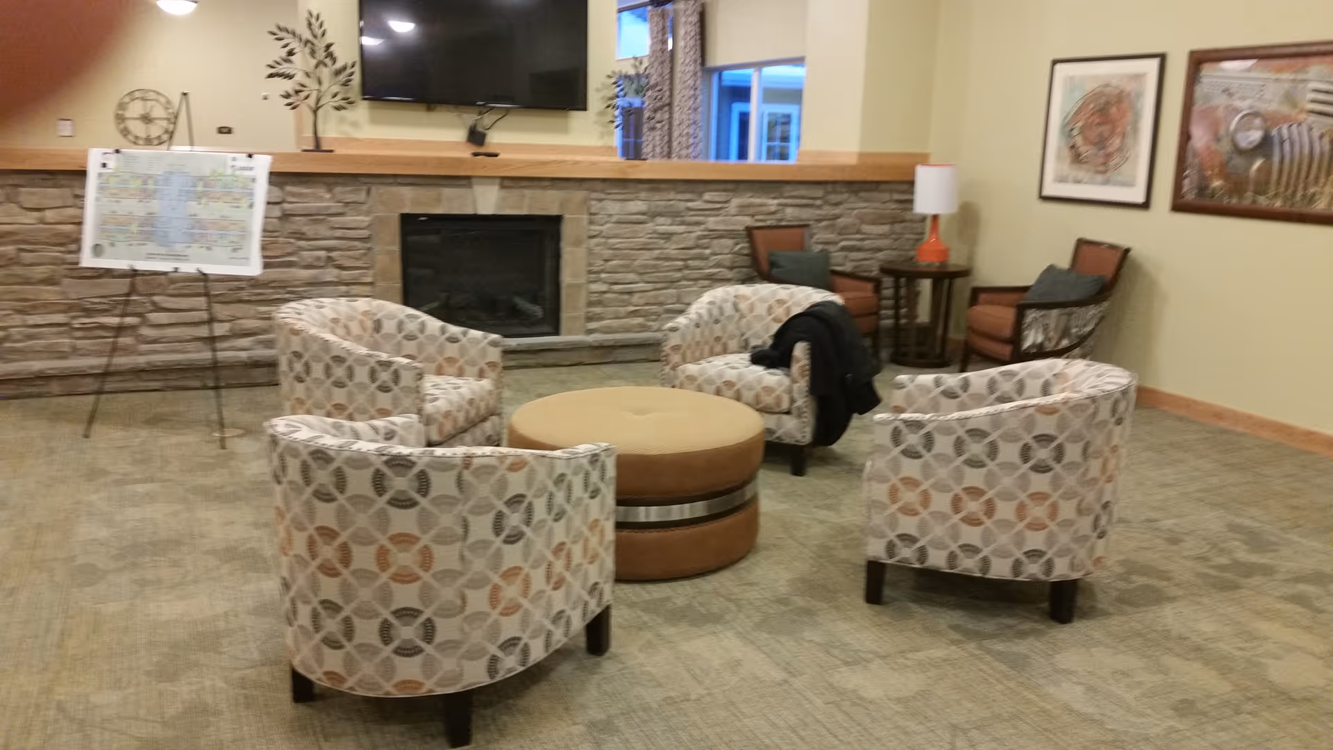 A cozy seating area in a senior living facility featuring four patterned armchairs arranged around a round ottoman. The room has a stone fireplace with a mounted flat-screen TV above it. There are two additional chairs with a small table and lamp in the corner, framed artwork on the walls, and a large window letting in natural light.