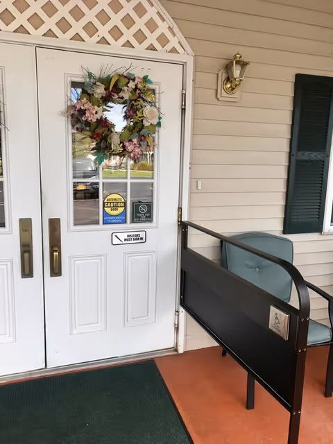 White double doors with glass panels, decorated with a floral wreath. Signs on the door indicate automatic caution door, no smoking, and visitors must sign in. To the right of the door is a green cushioned chair and a black metal bench with a handicap accessibility sign. The exterior wall is beige with a brass outdoor light fixture above the chair.