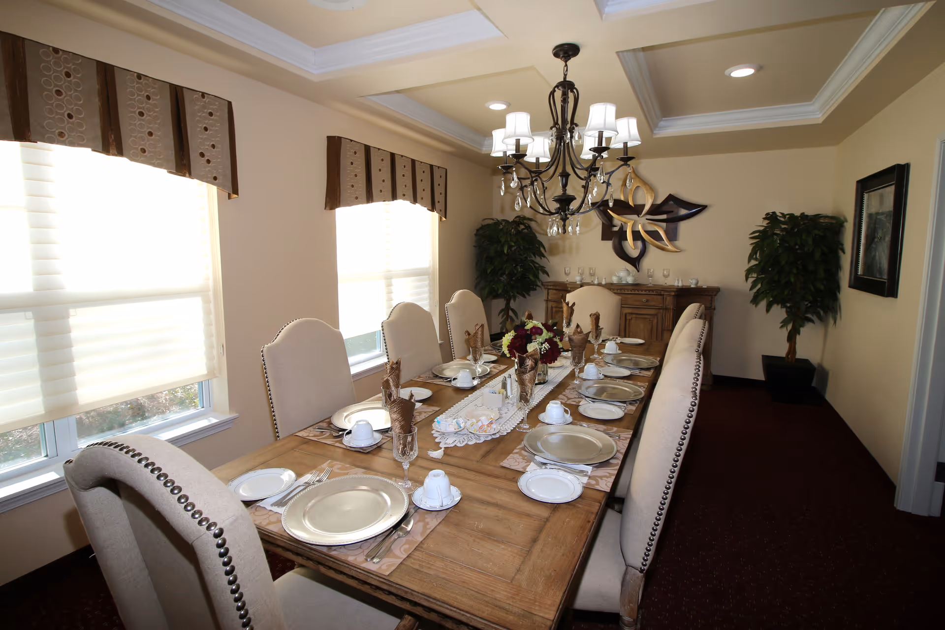 A formal dining room with a long wooden table set for eight people. The table is arranged with plates, cups, glasses, and napkins. There are upholstered chairs with nailhead trim around the table. Two large windows with blinds and valances let in natural light. A chandelier with multiple lights hangs above the table. The room has beige walls, a dark carpet, two potted plants, a wooden sideboard, and decorative wall art.