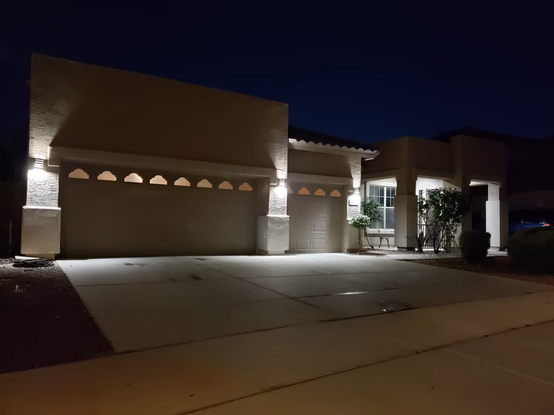 A single-story residential building at night with exterior lights illuminating the garage doors and entrance area. The driveway is empty and there are some bushes and small trees near the entrance.