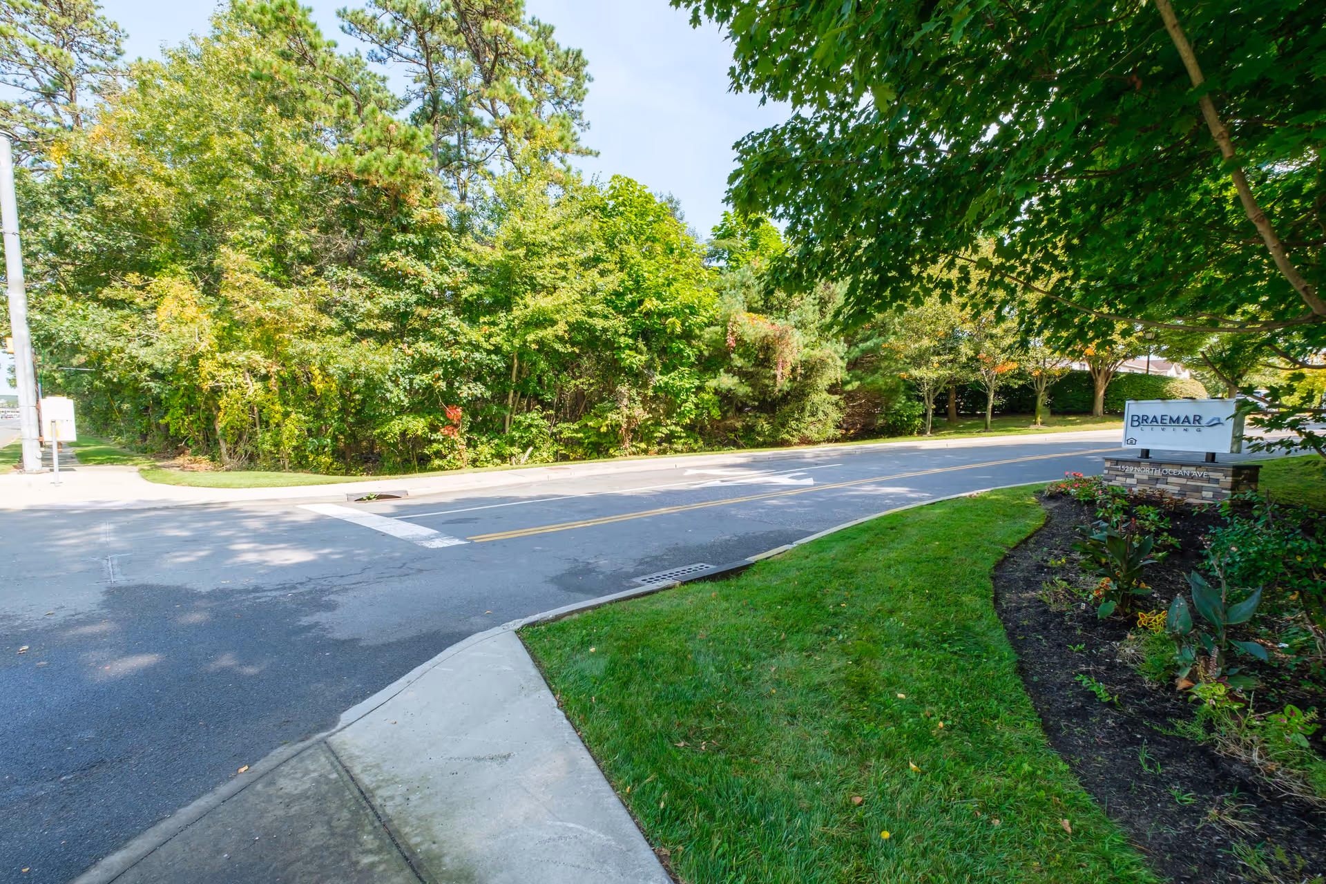 A paved road with a sidewalk and green grass on the side, surrounded by trees and bushes. There is a sign on the right side that reads 'Braemar at Medford' with some additional smaller text below it.