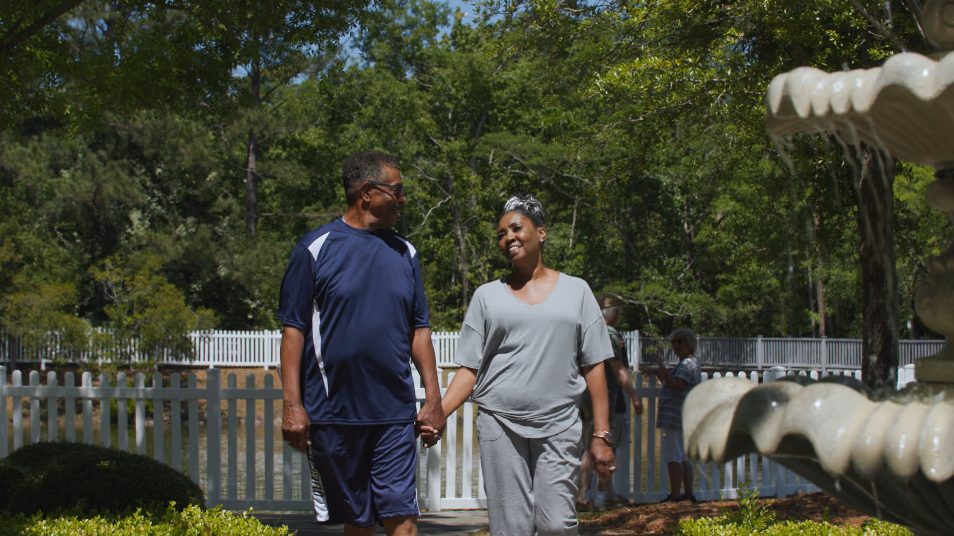 An older couple holding hands and walking outdoors near a white picket fence and a large water fountain, surrounded by green trees and bushes on a sunny day.