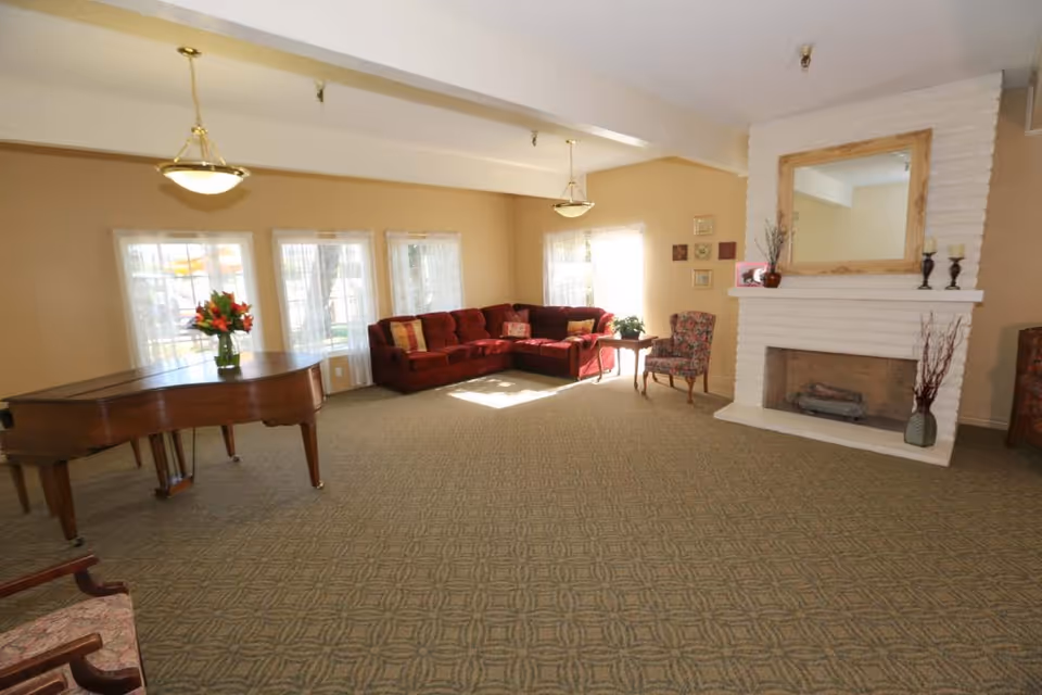 A spacious living room with beige walls and patterned carpet. The room features a red sectional sofa with colorful cushions, a floral armchair, a wooden side table with a plant, and a white brick fireplace with a large wooden-framed mirror above it. There is a grand piano with a vase of flowers on top near three windows with sheer white curtains, and two hanging ceiling lights.