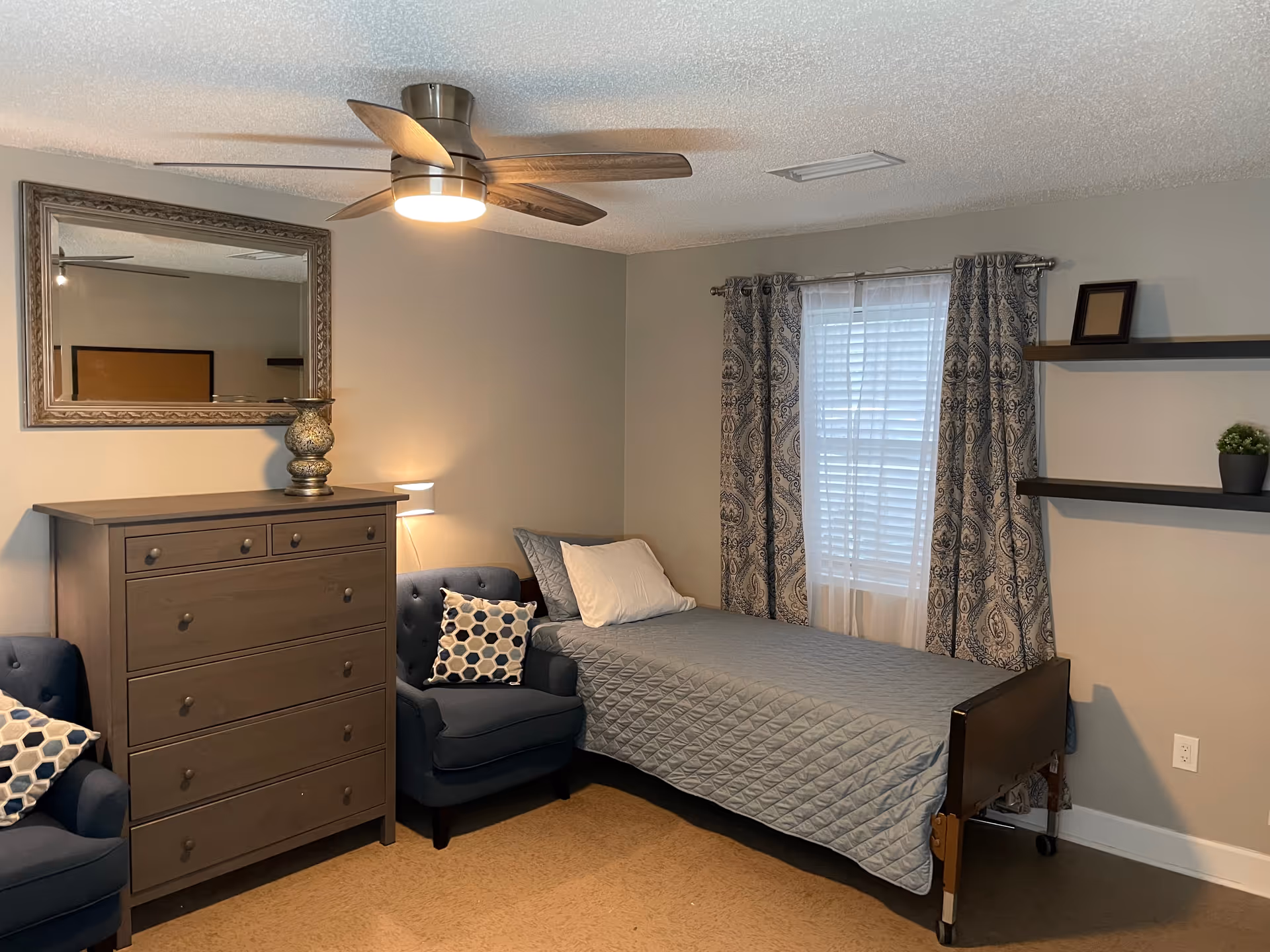 A neatly arranged bedroom with a single bed covered in a gray quilt, two blue armchairs with patterned cushions, a wooden dresser with a decorative vase on top, a large framed mirror above the dresser, patterned curtains on the window, and two floating shelves on the wall with small decorative items.