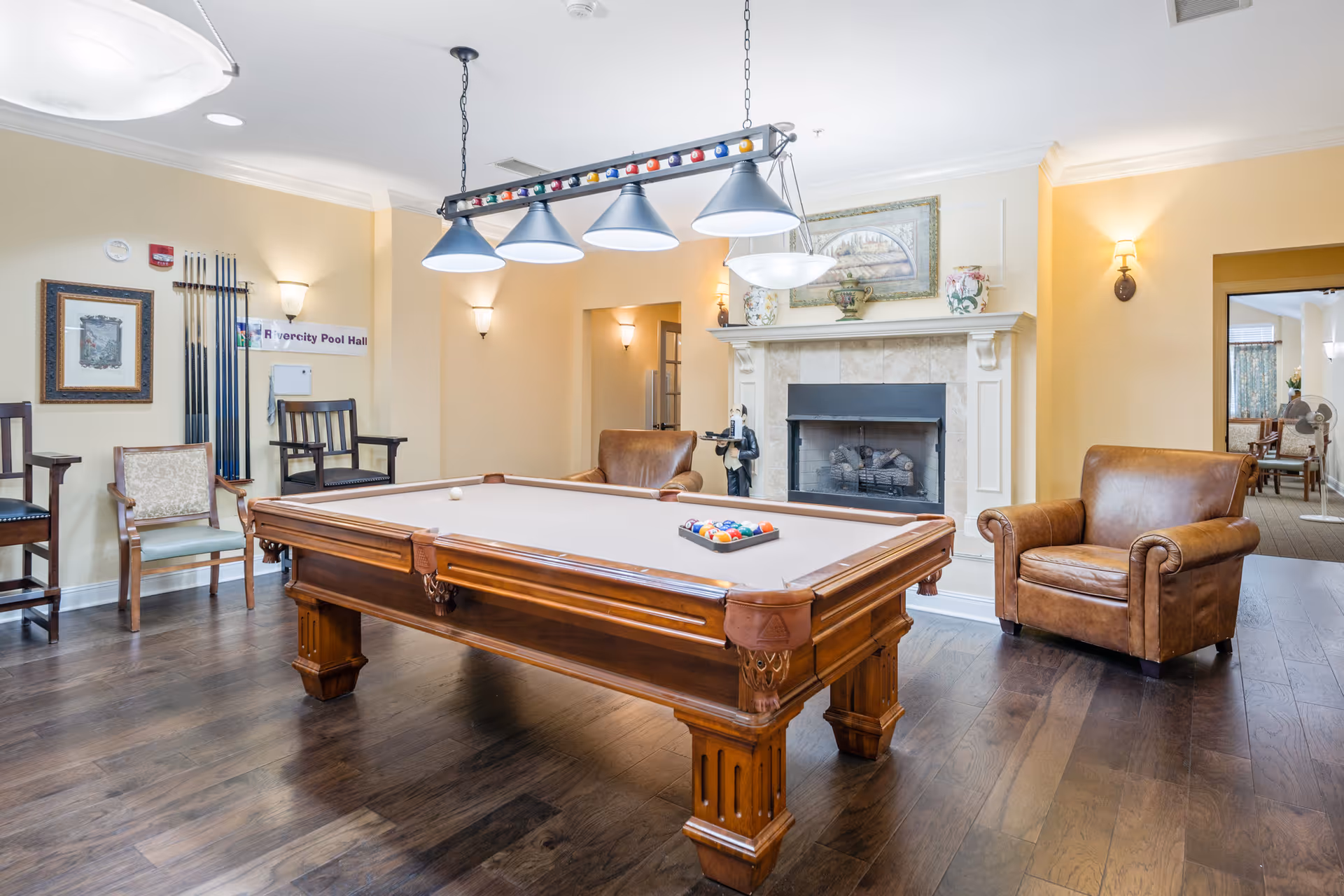 A well-lit recreational room featuring a pool table with balls arranged for a game, surrounded by various chairs including a leather armchair and wooden chairs. The room has a fireplace with decorative items on the mantel, a framed picture above it, and a rack with pool cues on the wall. The floor is wooden, and there are multiple light fixtures hanging from the ceiling and mounted on the walls.