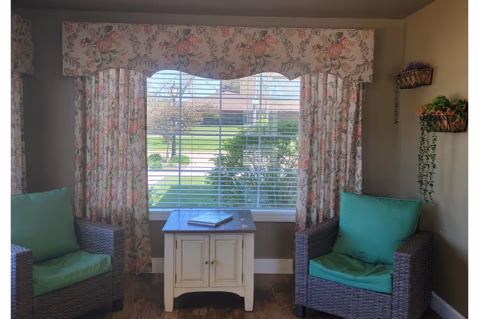 A cozy sitting area with two wicker armchairs featuring green cushions, positioned on either side of a small white wooden table with a book on top. Behind the table is a large window with white blinds and floral-patterned curtains and valance. The walls are beige, and there are two wall-mounted baskets with green and pink plants on the right side.