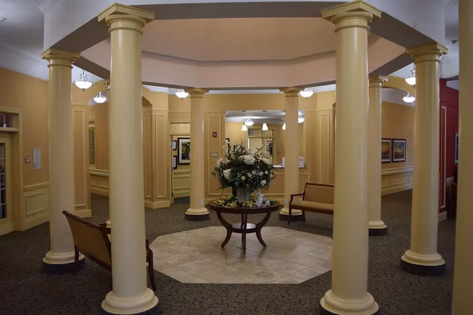 Interior view of a senior living facility lobby area with multiple cream-colored columns arranged in a circular pattern around a central table holding a large floral arrangement. The walls are painted in warm beige tones, and there are benches along the walls. Ceiling lights illuminate the space.