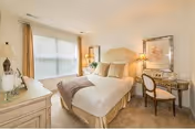 A cozy bedroom with a neatly made bed featuring beige and brown bedding. The room has a large window with beige curtains allowing natural light to fill the space. There is a white dresser with decorative items on the left side and a small desk with a chair and a lamp on the right side. The walls are light-colored, and there is framed artwork above the desk.