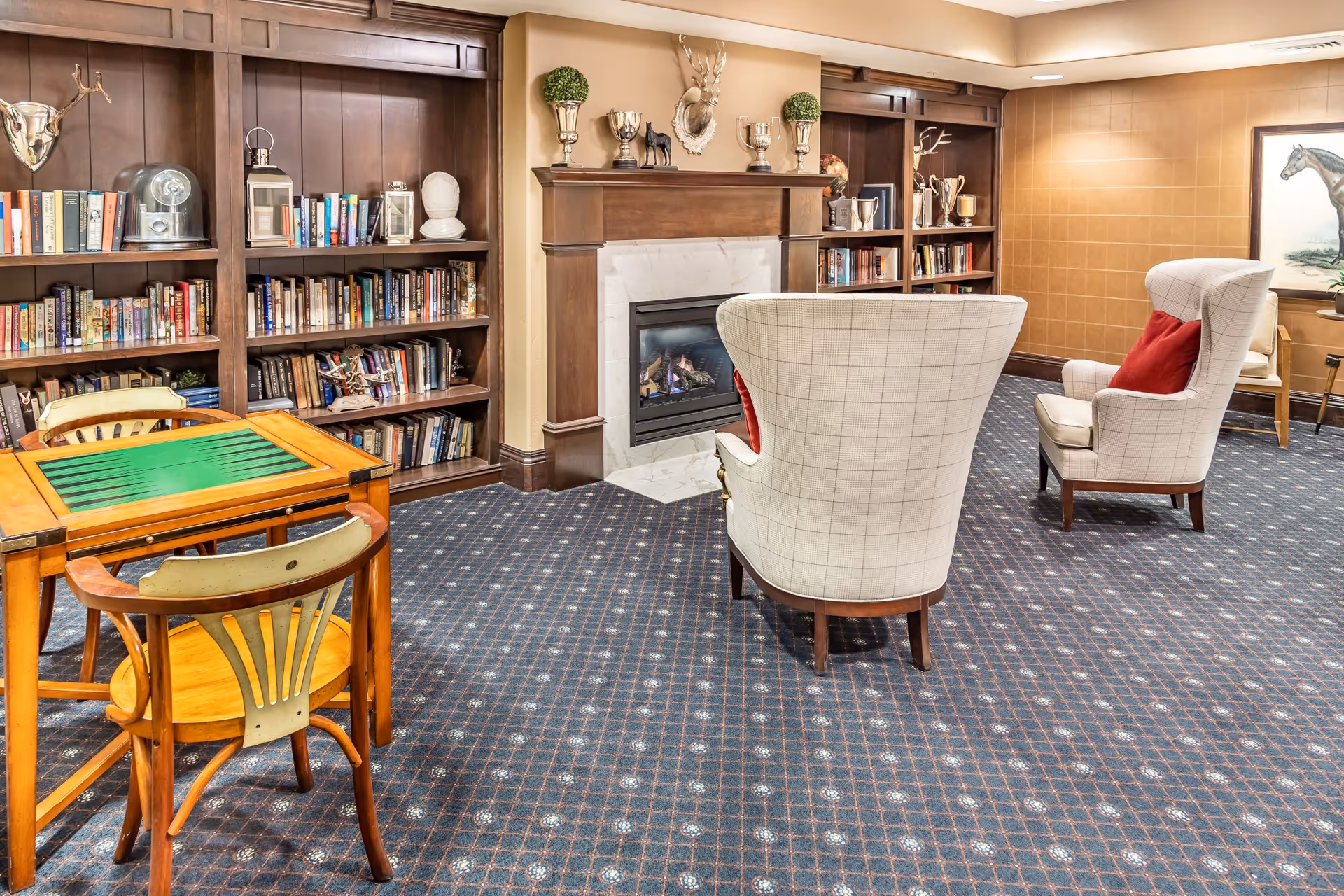 A cozy common area with a fireplace surrounded by wooden bookshelves filled with books and decorative items. Two upholstered armchairs with red cushions face the fireplace, and a small wooden table with two chairs is positioned nearby. The room has patterned carpet flooring and warm lighting, with a framed horse painting on the wall.