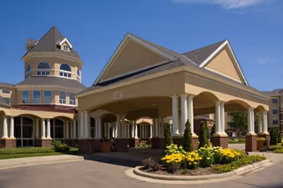 Exterior view of a senior living facility with a covered entrance supported by white columns, landscaped flower beds with yellow flowers, and a multi-story building with a turret under a clear blue sky.