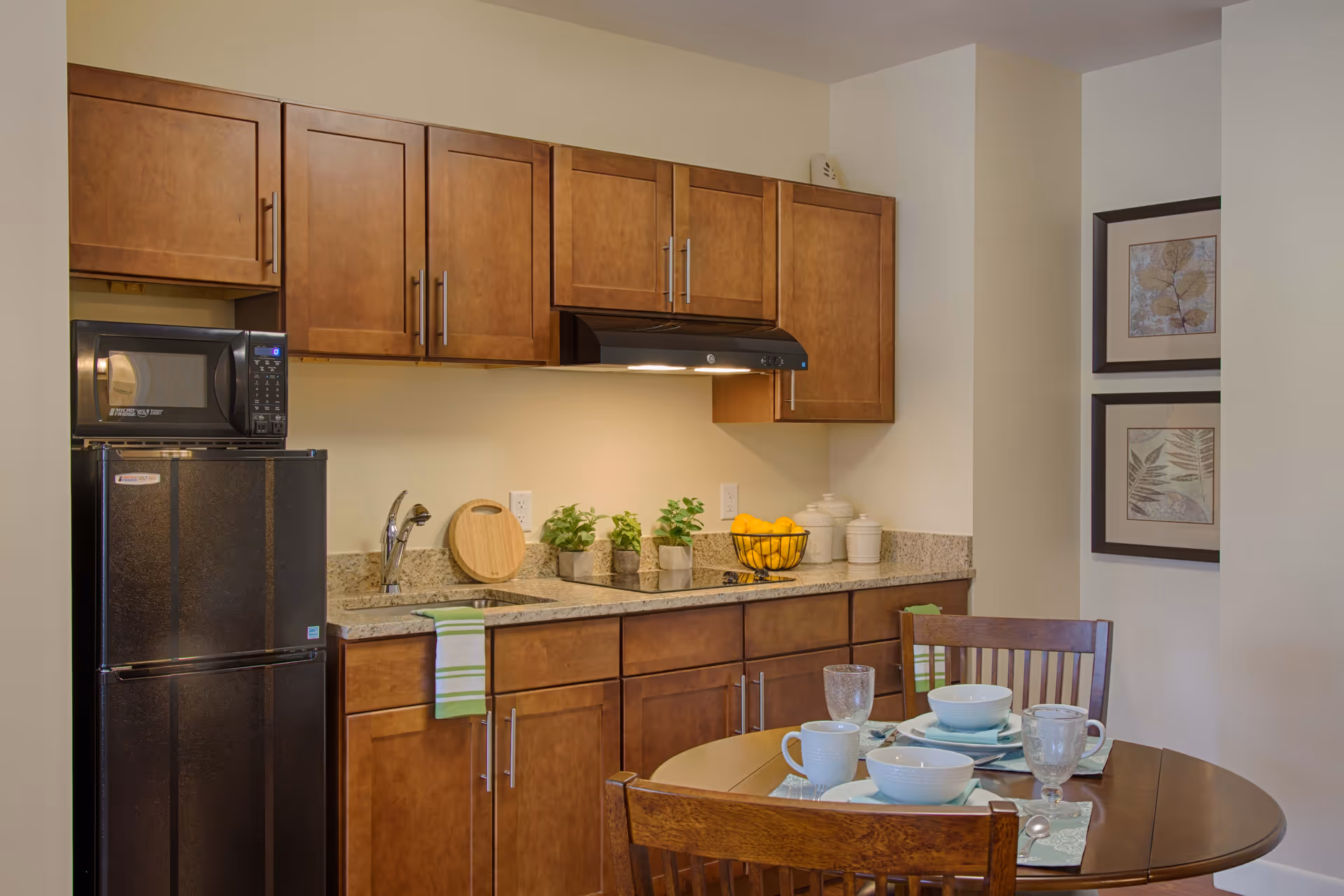 A small kitchen area with wooden cabinets, a black refrigerator with a microwave on top, a sink with a faucet, and a countertop decorated with small potted plants, a cutting board, a bowl of lemons, and canisters. In the foreground, there is a wooden dining table set with white bowls, cups, glasses, and silverware on placemats. Two framed botanical prints hang on the wall to the right.