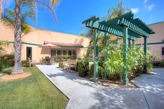 Outdoor courtyard area at a senior living facility with a green pergola surrounded by plants, benches, palm trees, and a building with windows and an umbrella-covered table in the background under a clear blue sky.