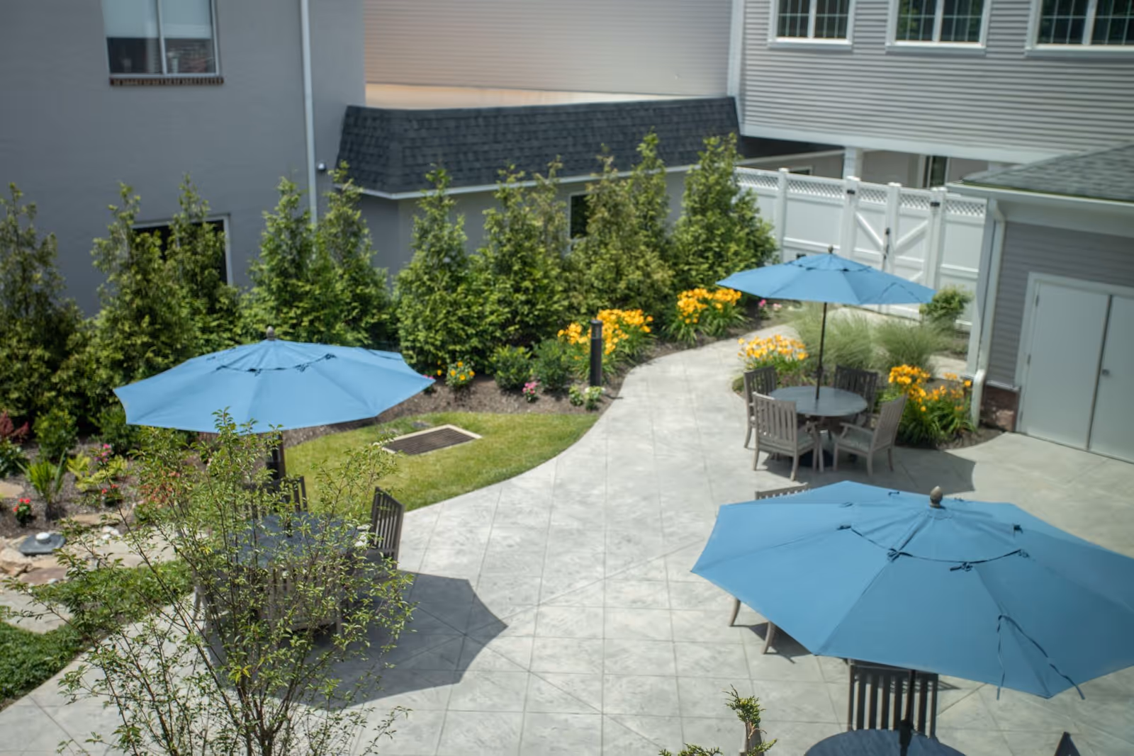 Outdoor patio area with several round tables and chairs, each shaded by large blue umbrellas. The patio is surrounded by greenery including bushes, flowers, and trees, with buildings visible in the background.