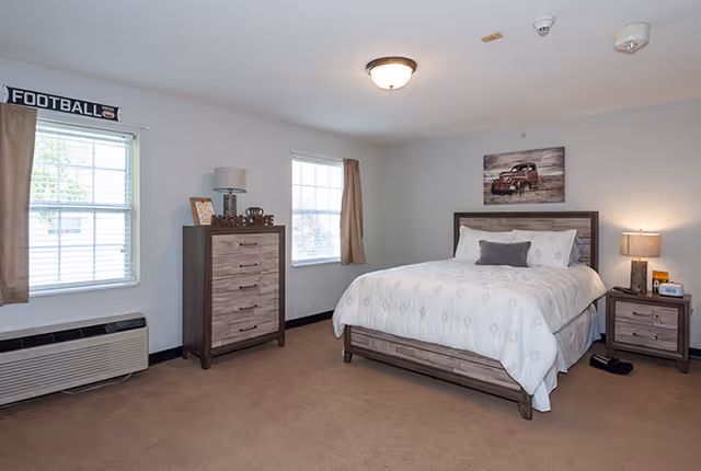 A spacious bedroom in an assisted living community featuring a large bed with white bedding and a gray accent pillow, a wooden nightstand with a lamp and clock, a matching wooden dresser with decorative items, two windows with brown curtains, and a wall decoration of a vintage car above the bed.