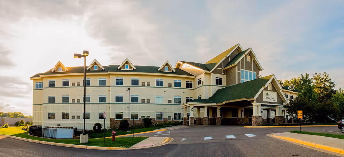 Front exterior of a multi-story assisted living building with a covered porte-cochère, landscaped grounds, and a driveway.