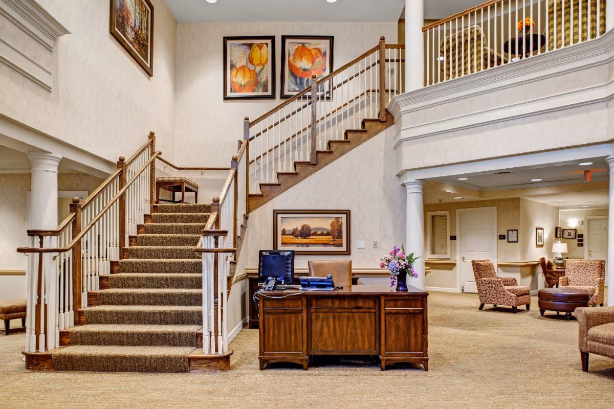 Lobby of a senior living facility with a central carpeted staircase, wooden reception desk, and seating area.