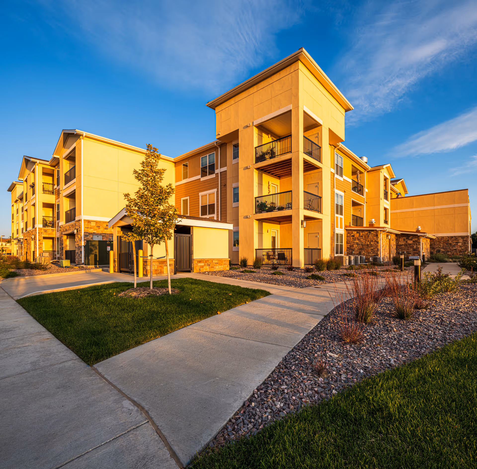 Sunlit three-story residential building with balconies, landscaped walkways, and a small tree under a blue sky.