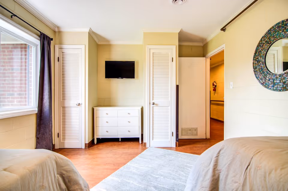 A bright bedroom with two beds partially visible on either side, a white dresser with drawers in the center against a beige wall, a flat-screen TV mounted above the dresser, two white louvered closet doors on either side of the dresser, a large window with dark curtains on the left, a round decorative mirror on the right wall, and a doorway leading to a hallway with a fire extinguisher visible.