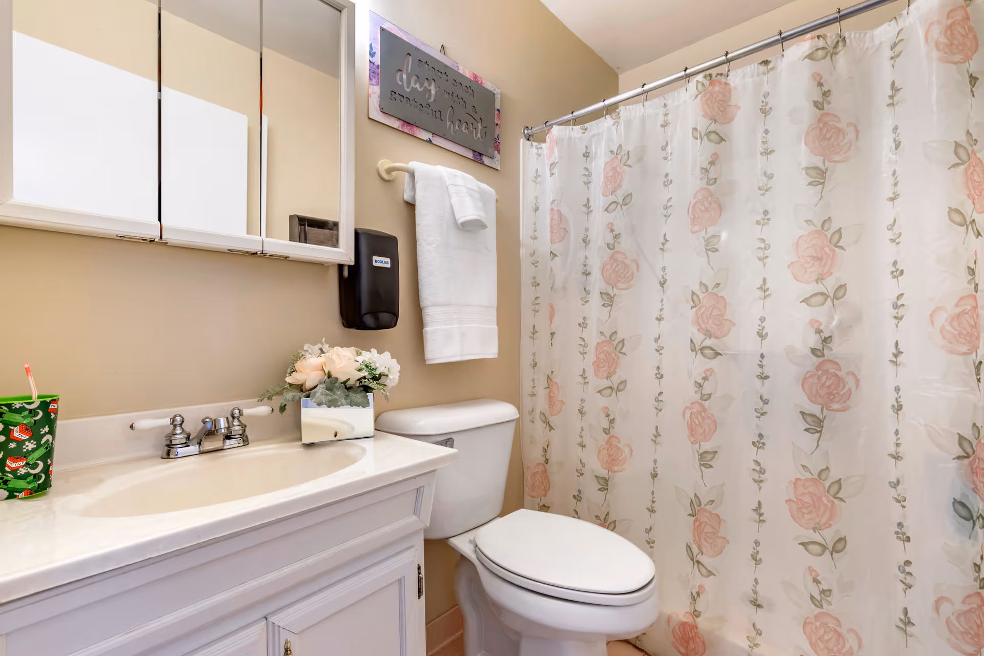 A bathroom with a white vanity and sink, a toilet, and a shower with a floral-patterned curtain. Above the toilet, there is a towel rack with a white towel and a decorative sign on the wall. A green cup with a straw is on the vanity next to a small flower arrangement.