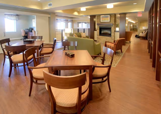 A spacious senior living common area featuring wooden tables and chairs in the foreground, a green sofa and armchairs arranged around a stone fireplace, and a hallway with additional seating and bookshelves along the right side. Large windows provide natural light to the room.