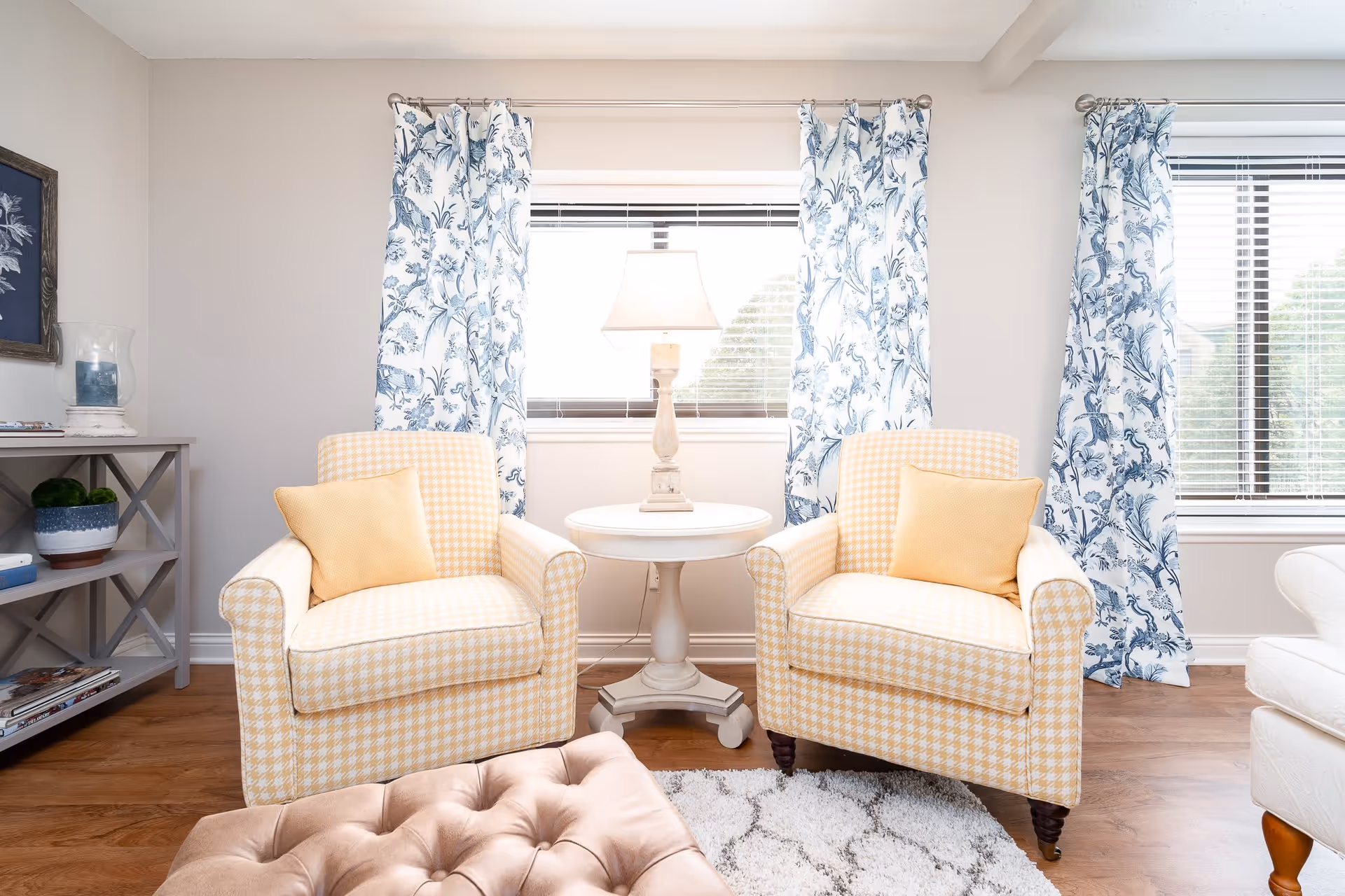 A bright living room area with two yellow and white checkered armchairs, each with a yellow pillow. Between the chairs is a small round white table with a lamp on it. The room has two windows with blue and white floral curtains and wooden flooring. A beige tufted ottoman and a gray shelf with decorative items are also visible.