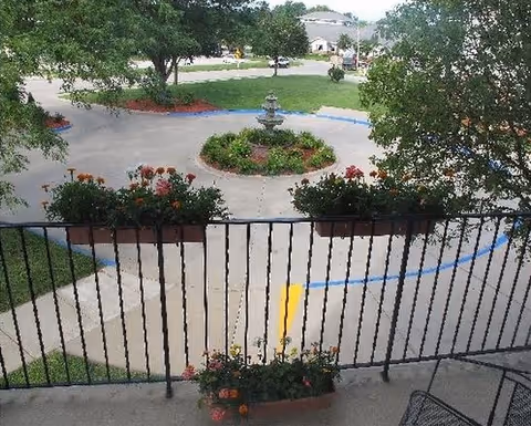 View from a balcony over a circular driveway with a central fountain, flower planters on the railing, and trees and lawns beyond.