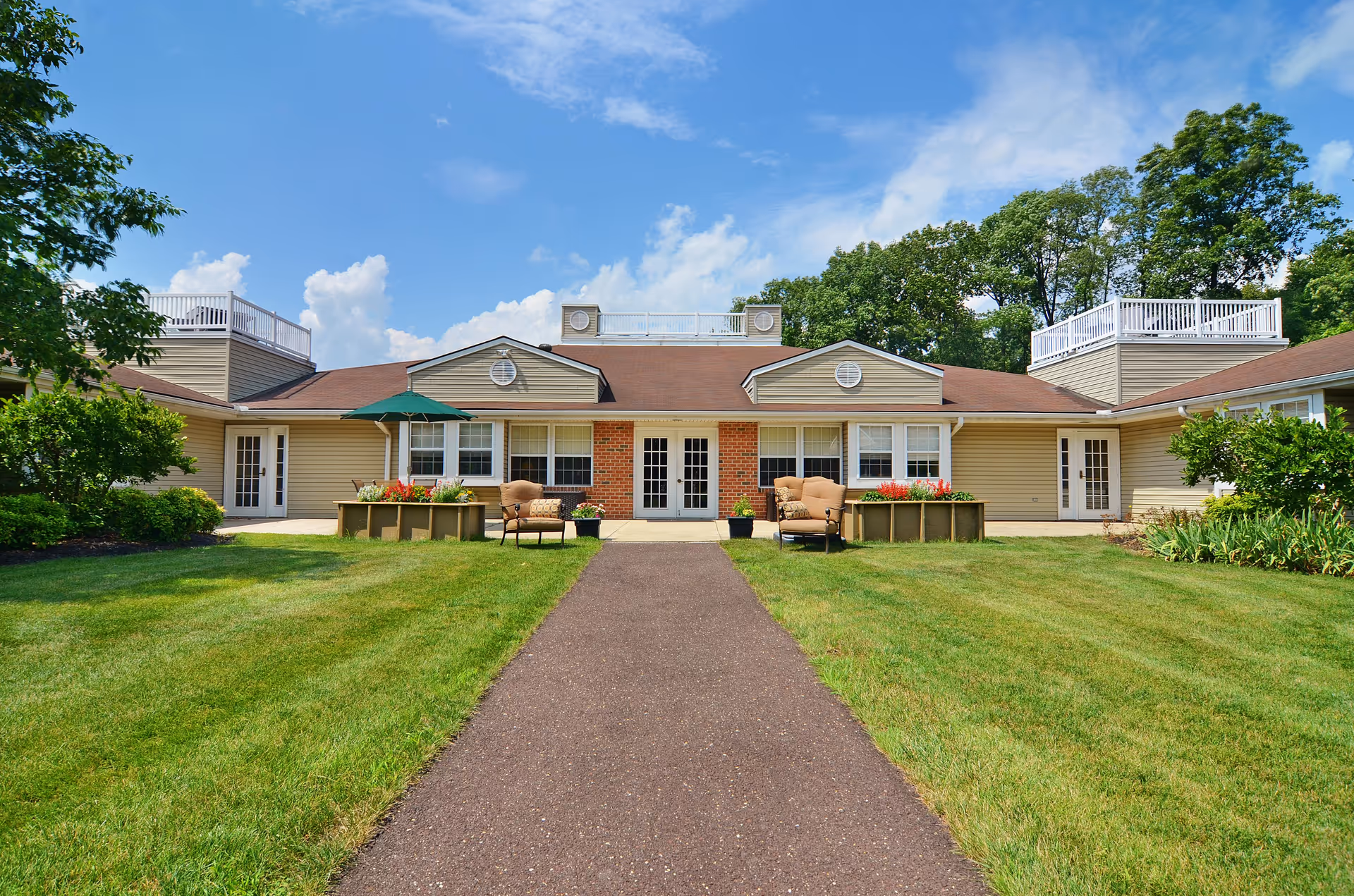 Exterior view of a single-story building with beige siding and a brown roof, featuring a central entrance with double doors and windows on either side. There are two seating areas with cushioned chairs and a green umbrella on a paved pathway leading to the entrance. The building is surrounded by well-maintained green lawns, flower beds, and trees under a blue sky with scattered clouds.