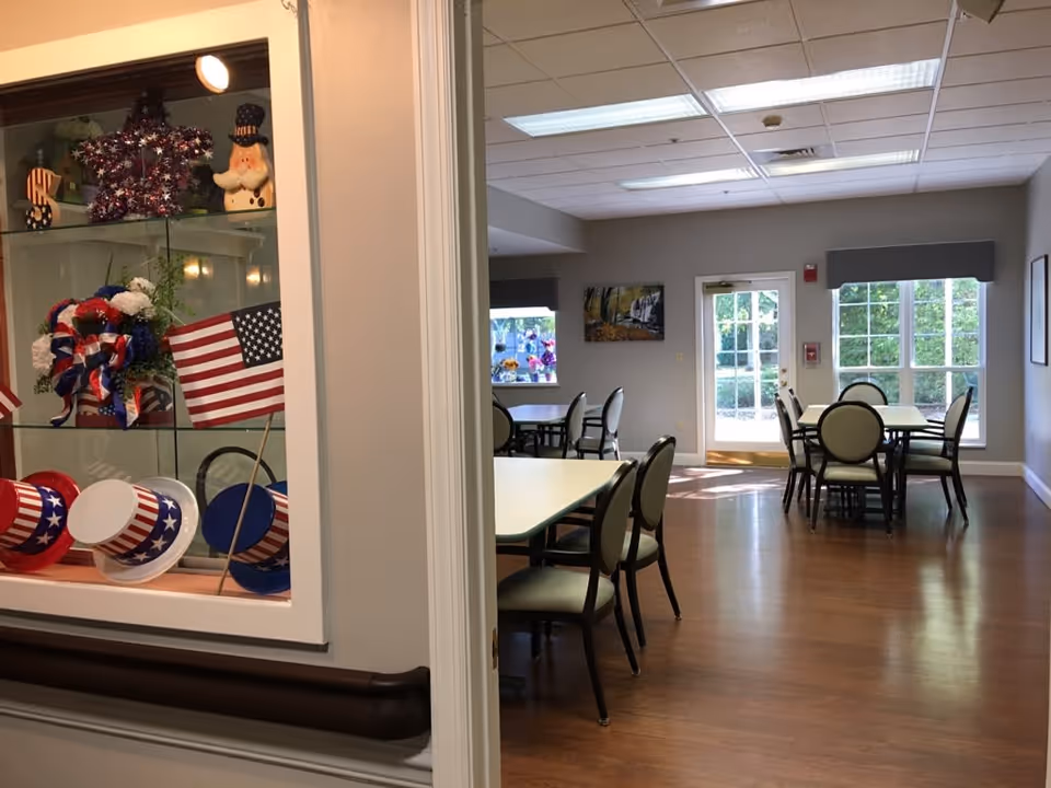 A bright dining/activity room with tables and chairs seen through a doorway next to a glass display case filled with patriotic decorations.