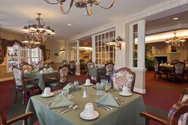 Elegant dining room with tables draped in green tablecloths, formal place settings, floral-upholstered chairs, and chandeliers.