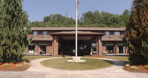 Front exterior view of a two-story brick building with a covered entrance and a flagpole in the center of a circular driveway. There are trees and shrubs around the building and a person walking near the entrance.