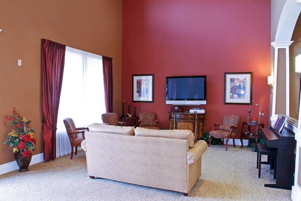 A cozy living room with a beige sofa facing a wall-mounted flat screen TV. The walls are painted in warm tones of brown and red. There are four upholstered chairs arranged around the sofa, two framed pictures on the red wall, a wooden cabinet under the TV, a piano on the right side, and a large window with red curtains on the left. A floral arrangement is placed in the corner near the window.