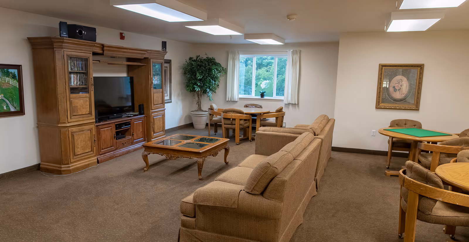 A communal lounge with sofas facing a large wooden TV entertainment center, a coffee table, and game tables by a window.