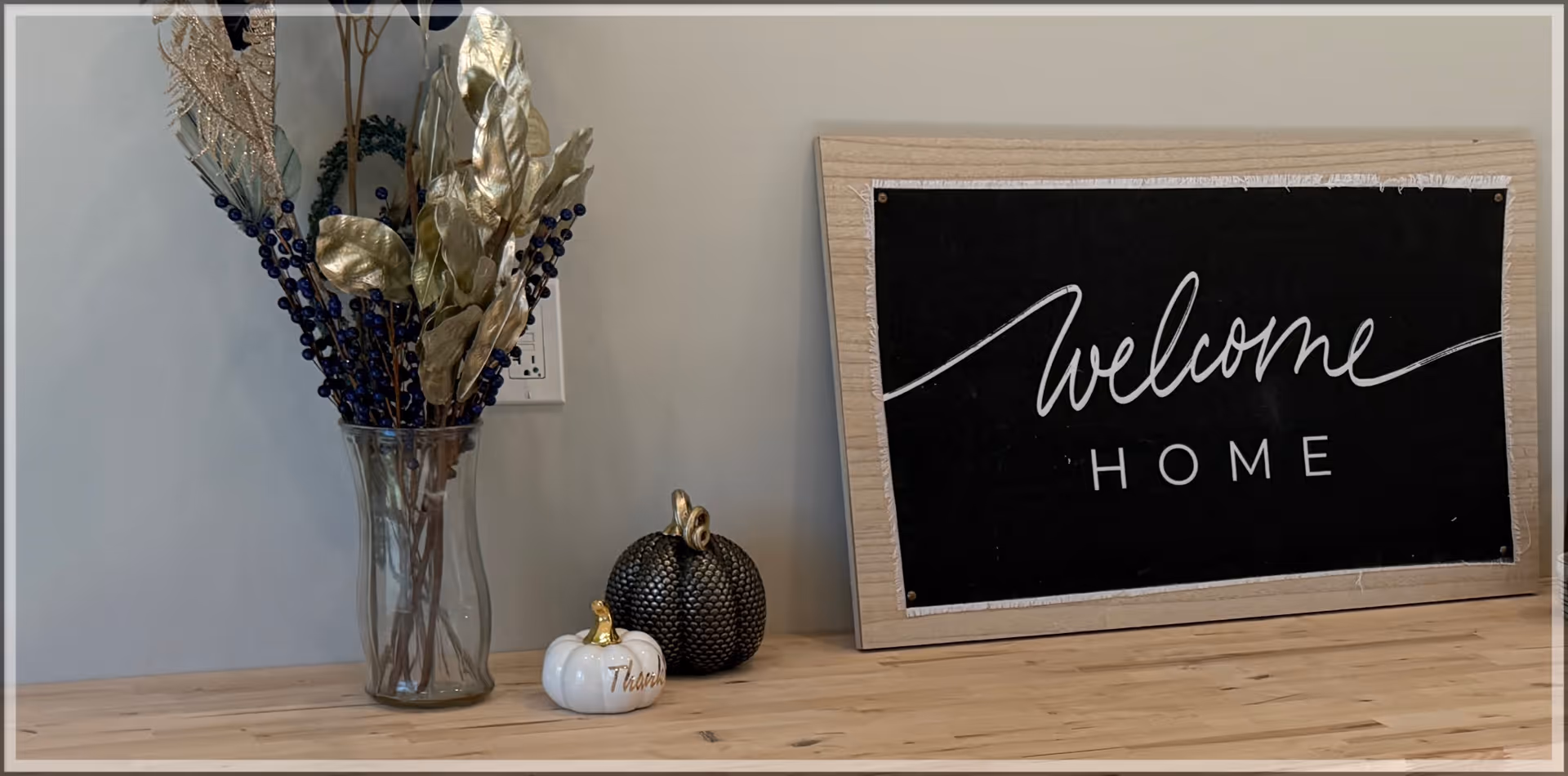 A wooden surface with a glass vase holding decorative dried plants, two small ornamental pumpkins, and a framed blackboard sign that reads 'welcome HOME'.