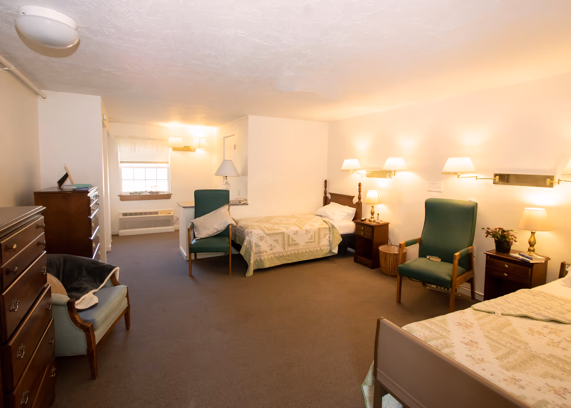 A spacious bedroom in a senior living facility with two single beds, each with a nightstand and lamp. There are two green armchairs, a wooden dresser, and a small window letting in natural light. The room has beige walls and carpeted floor, with soft lighting from wall-mounted lamps.