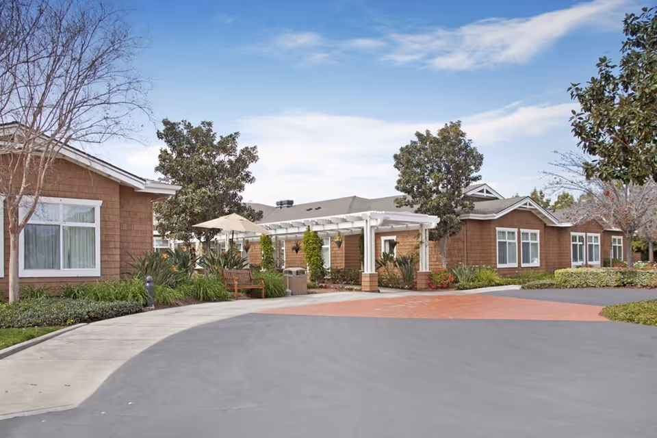 Front exterior of a single-story senior living building with a covered entrance, landscaping, benches, and trees.