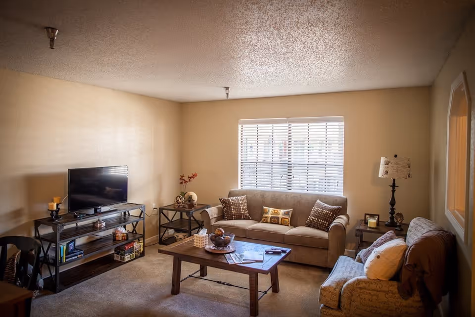 A cozy living room with a beige couch adorned with patterned pillows, a patterned armchair with a white pillow and brown throw, a wooden coffee table with decorative items, a TV on a metal and wood stand, and a window with white blinds letting in natural light.